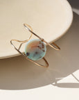 The siren cuff features a Terra Agate Gemstone wire wrapped to a 14k gold fill wide cuff. The cuff is displayed against a cream plate in the sunlight. 