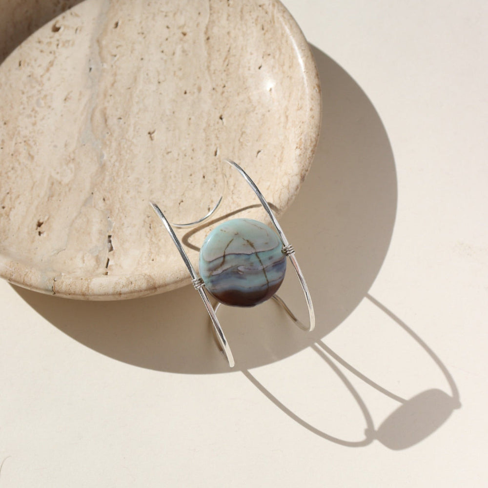 The siren cuff features a Terra Agate Gemstone wire wrapped to a sterling silver wide cuff. The cuff is displayed against a cream plate in the sunlight. 