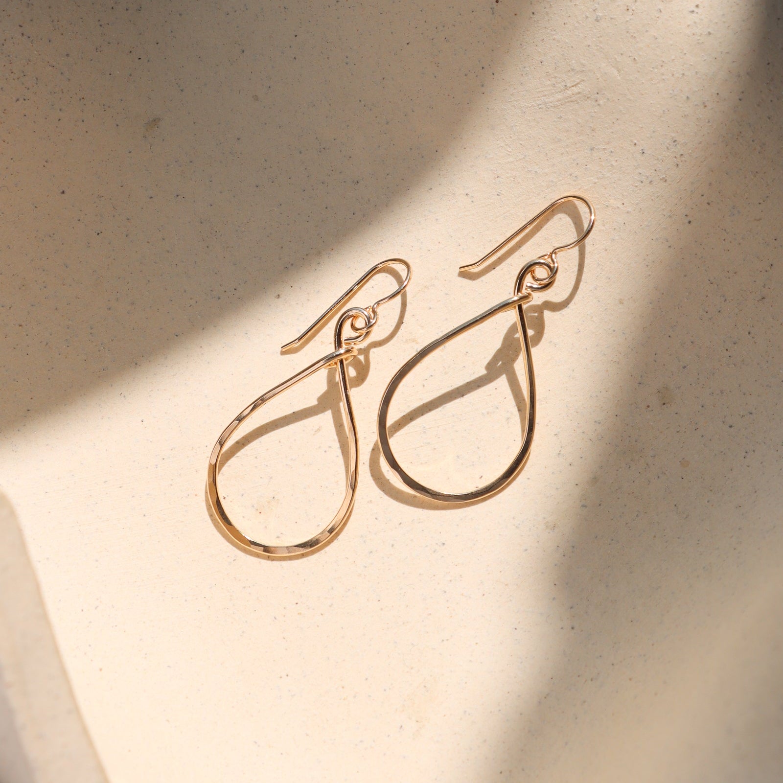 A pair of gold-toned teardrop dangle earrings with a polished finish, displayed on a textured beige surface with soft diagonal shadows.