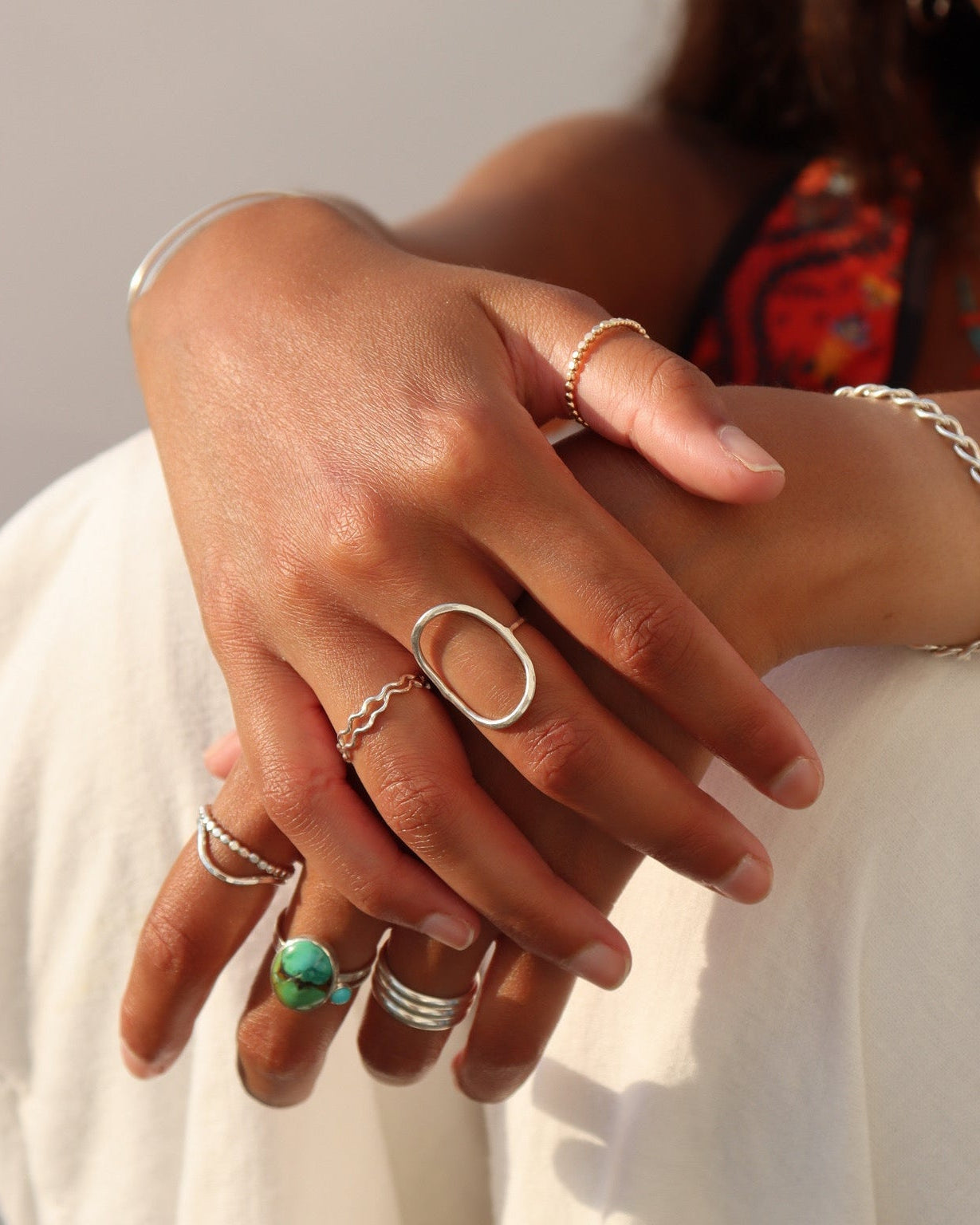 Close-up lifestyle image of a person wearing layered sterling silver rings, including a bold oval statement ring, wavy stacking ring, beaded bands, and a natural turquoise statement ring. A chunky silver chain bracelet and slim cuff complete the look, photographed in warm golden light against a soft neutral background.