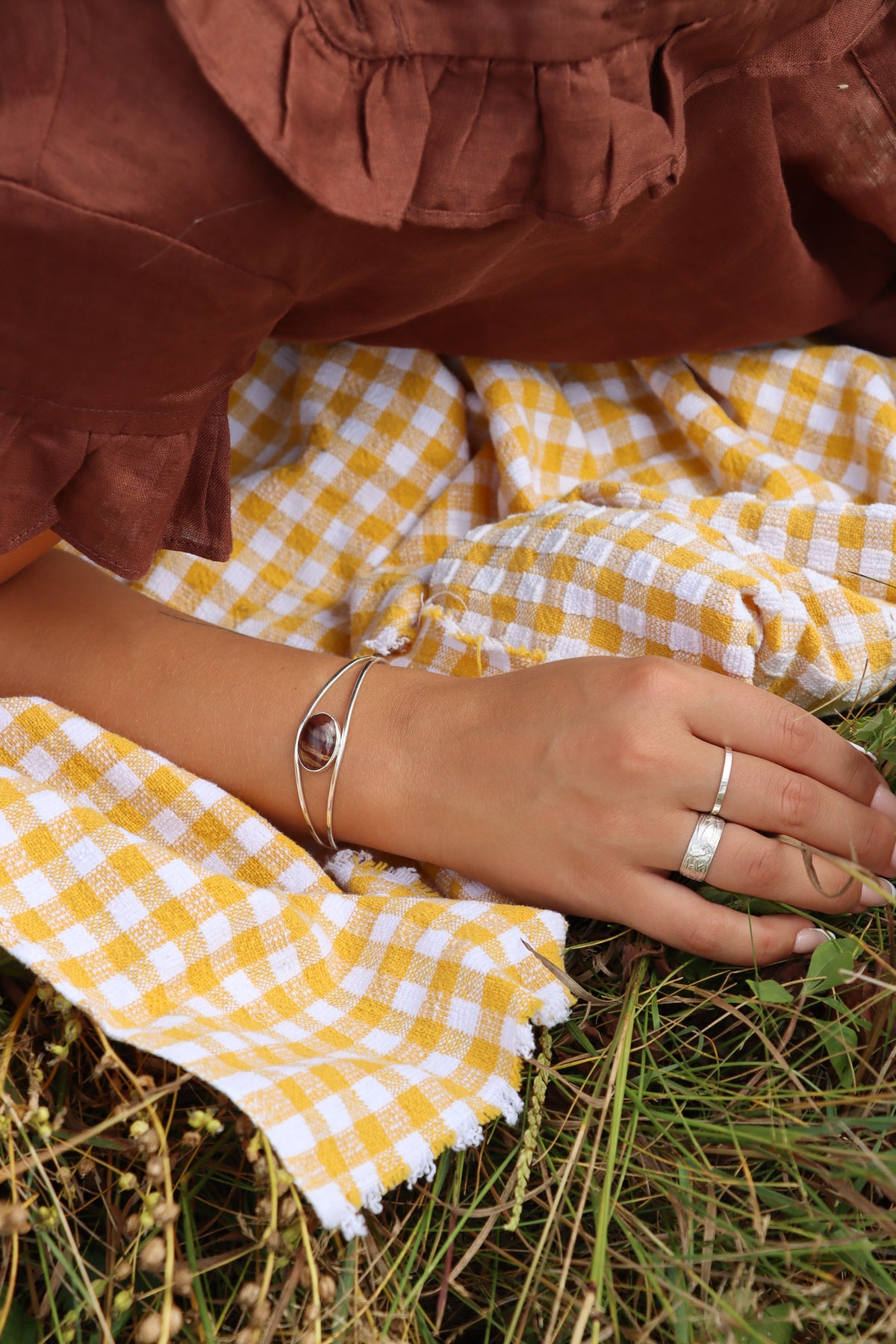 Silver open cuff bracelet with an oval banded red and brown stone centerpiece, worn on the wrist and styled with silver rings against yellow gingham fabric.