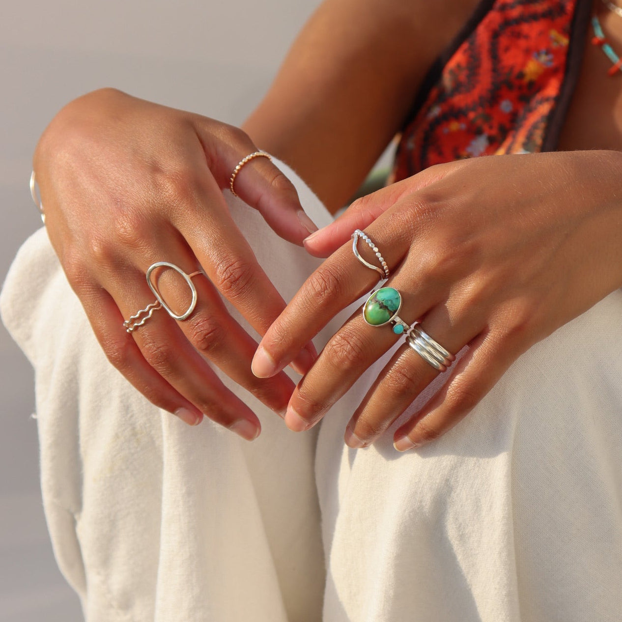 A young woman wearing white linen pants and an orange patterned top, seated in warm sunlight with hands resting on her knees to highlight an array of sterling silver stackable rings, including a turquoise stone bezel ring, a wavy silver band, and an open oval statement ring