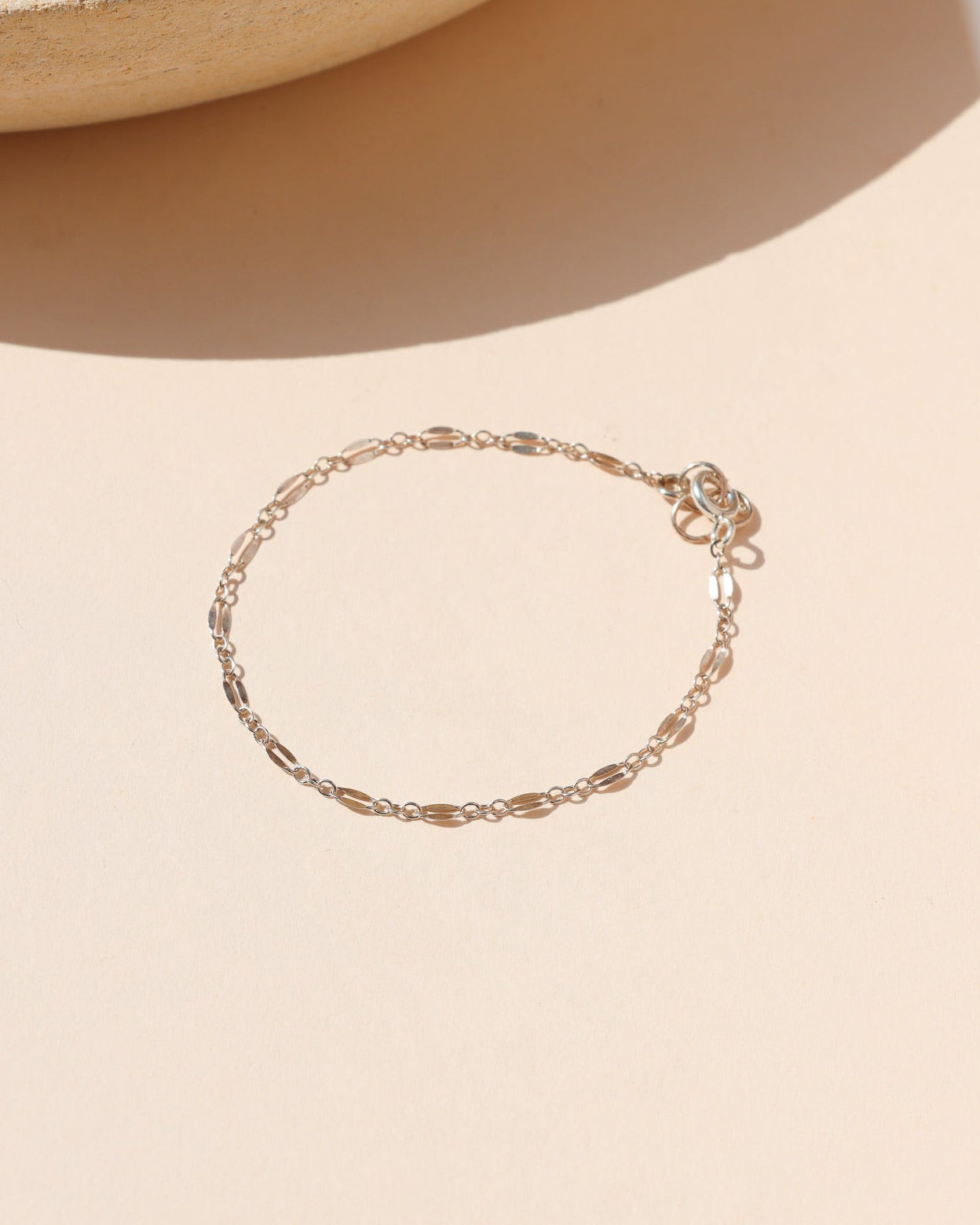 A delicate silver chain bracelet arranged flat in a circular shape on a light beige surface. The fine chain features alternating flattened oval links and a spring ring clasp, set against a warm background with a ceramic tray edge casting soft shadows.