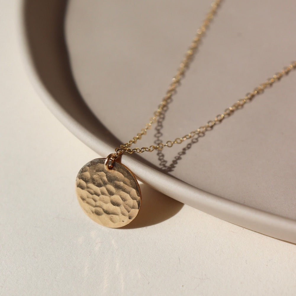 14k gold fill sun drop necklace laid on a tan plate in the sunlight. This necklace features our simple chain met by a hammered coin, making it look like the moon.