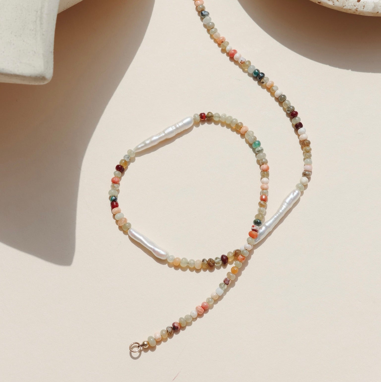 Smooth beaded necklace featuring pink, red, green, orange, teal, and cream Mexican Opals, laid organically on a beige background in the sunlight. 