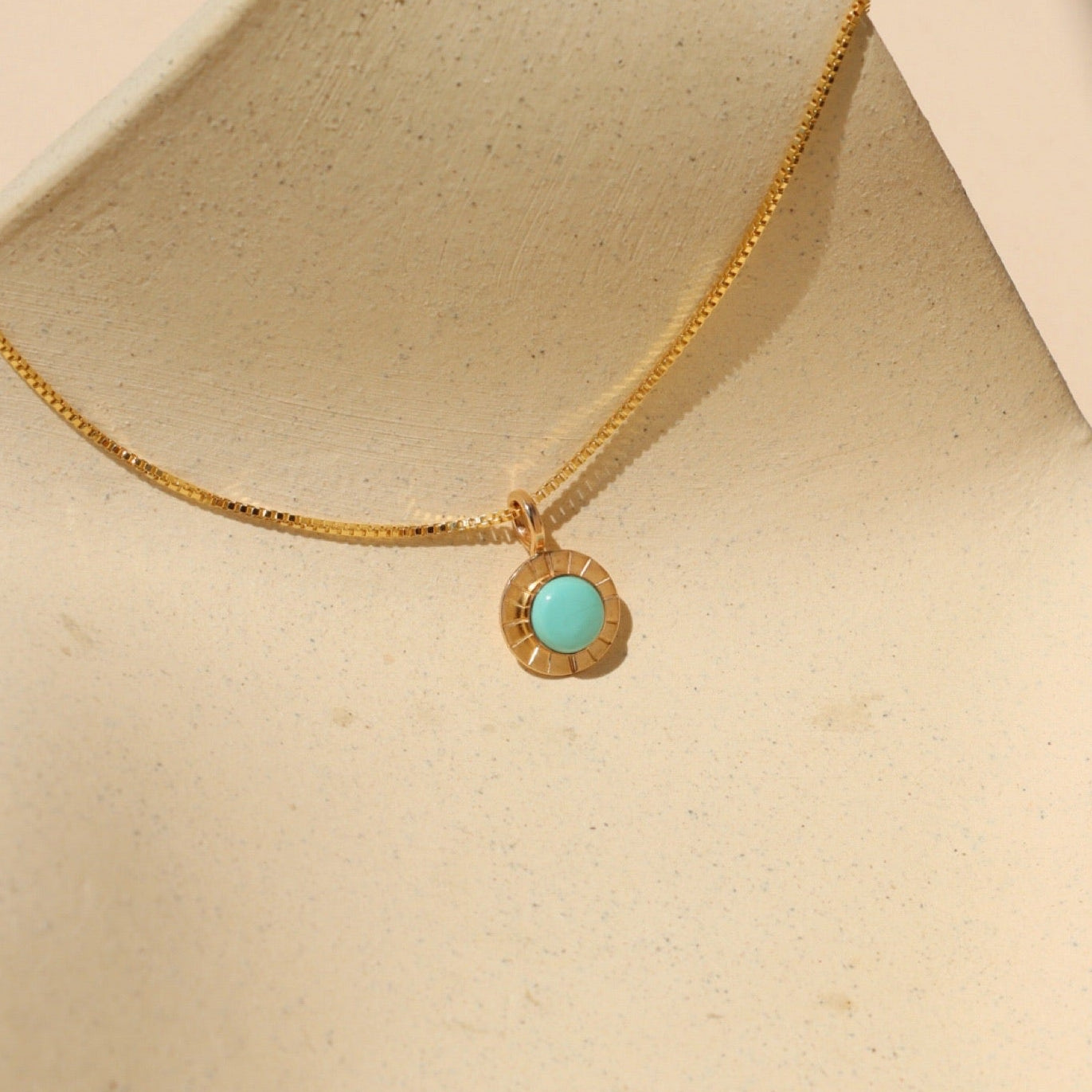 Gold necklace with a delicate box chain and a small round pendant featuring a bright turquoise stone set in a textured gold bezel, displayed on a sculptural cream ceramic stand in warm natural light.
