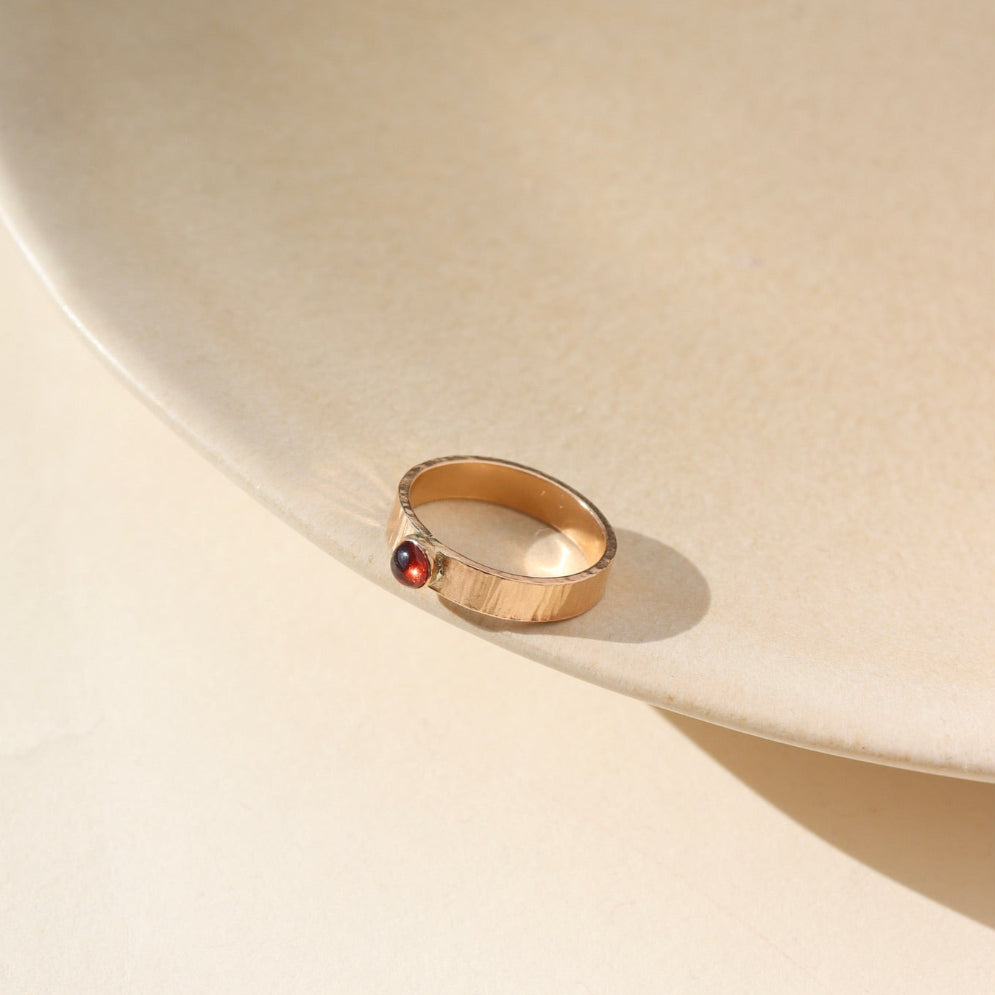 A ridged textured 14k gold fill band with a 4mm red garnet gemstone set in the middle. Ring is displayed on a tan dish. 