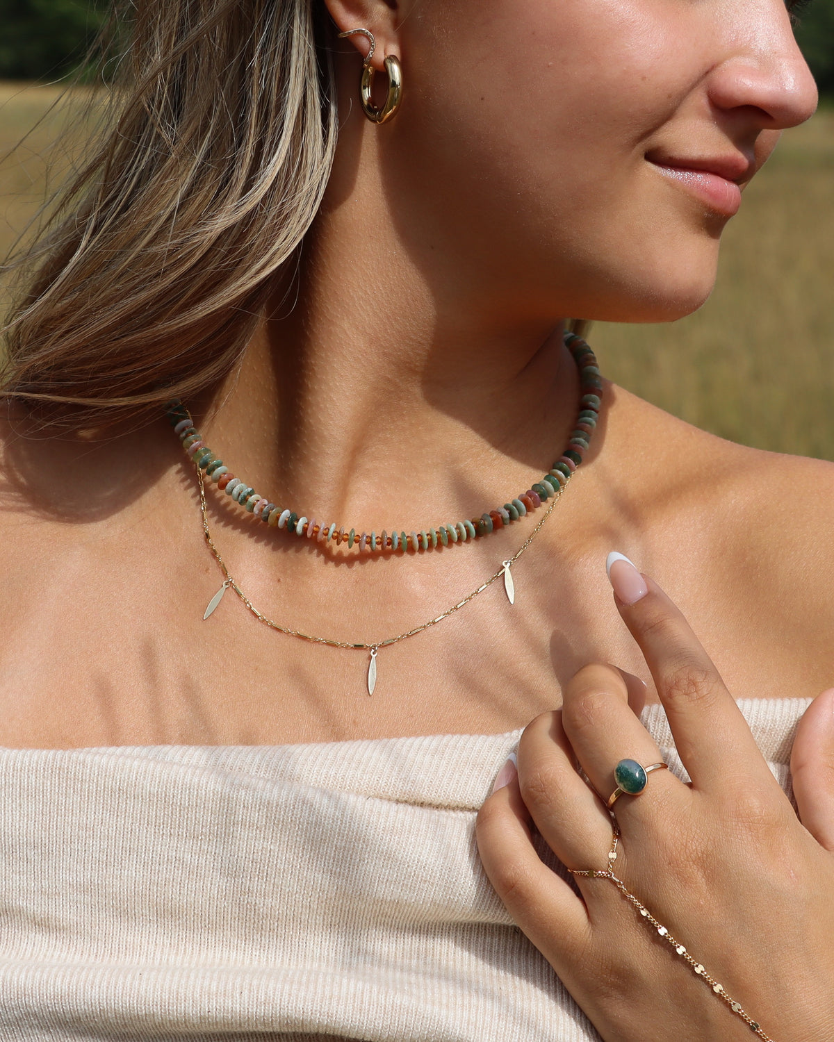 Gold charm necklace with three elongated leaf-shaped dangles, layered beneath a multicolor gemstone beaded necklace.