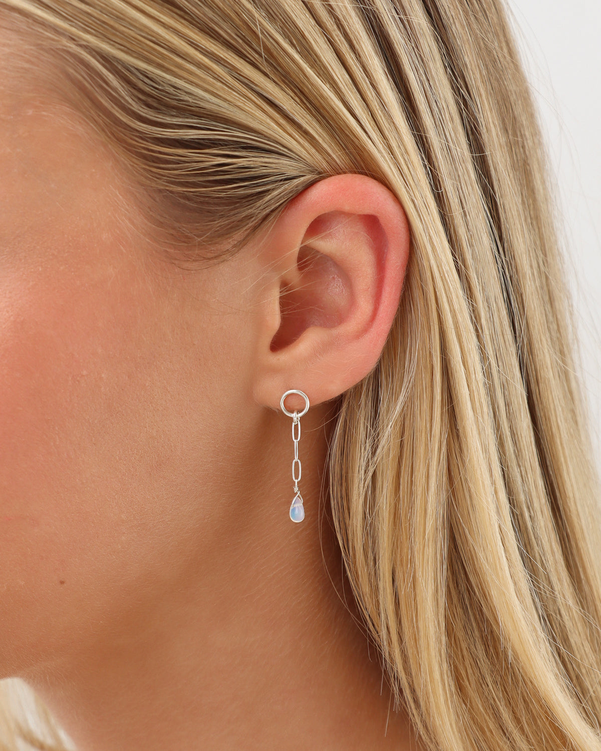 Close-up of a model wearing a silver opal drop earring featuring a circular stud, delicate elongated chain links, and a small iridescent teardrop opal pendant against blonde hair.