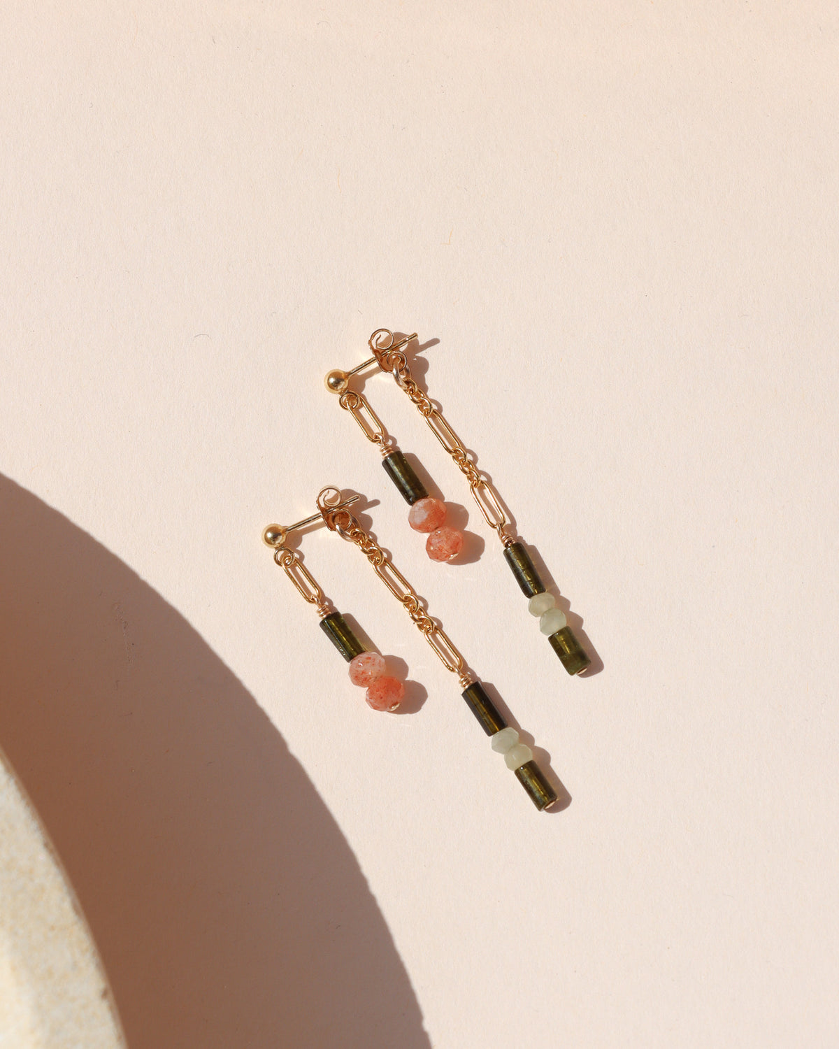 Gold-filled mismatched drop earrings with delicate chain links and green, white, and peach gemstone beads, displayed in warm sunlight.