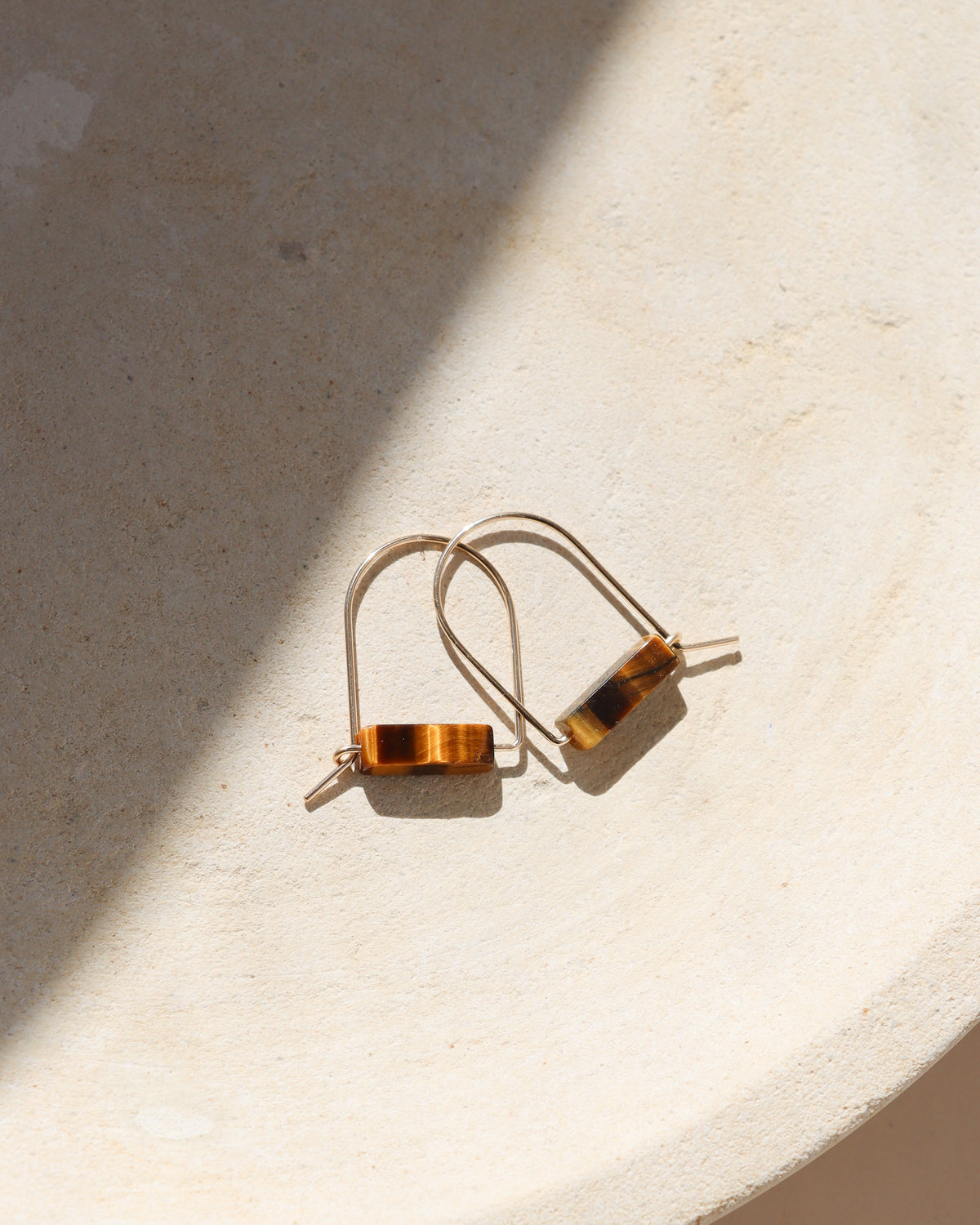 gold wire earrings featuring warm golden-brown Tiger’s Eye gemstone beads, arranged on a textured cream surface with soft shadows and natural sunlight.