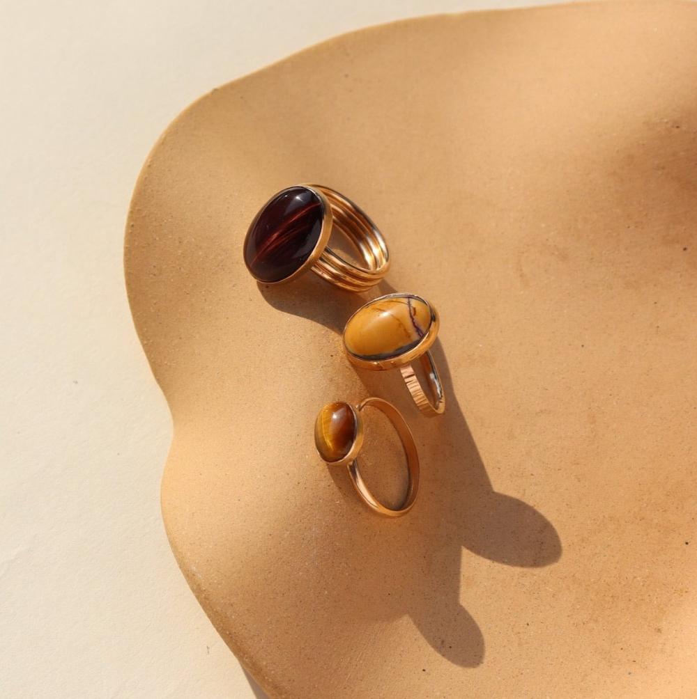 An amber-colored Tiger Eye gemstone on a horizontal bezel on a 14k gold fill band, photographed next to two other rings with larger mookite gemstones