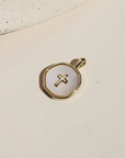 Round mother of pearl charm with a gold cross detail and gold-plated border, photographed on a neutral surface in warm natural light.