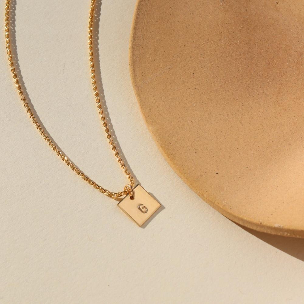 A small gold chain with a 3/8 x 3/8 square pendant stamped with a "G". Necklace is displayed on a cream backdrop. 