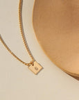 A small gold chain with a 3/8 x 3/8 square pendant stamped with a "G". Necklace is displayed on a cream backdrop. 