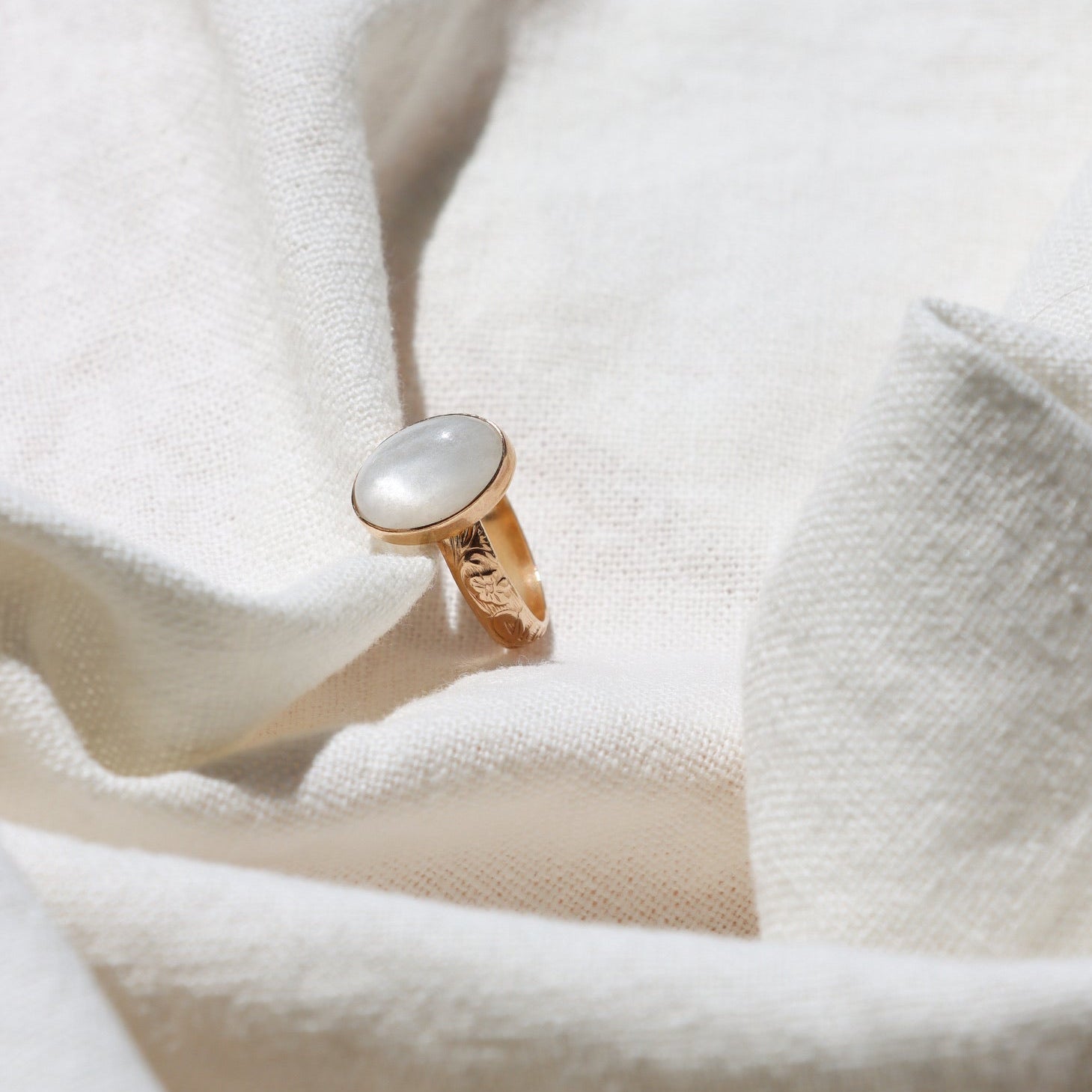 Close-up product photo of the White Lotus Ring, featuring an oval white moonstone set on a hand-engraved 14k gold fill floral band, displayed on soft ivory linen in warm natural sunlight.