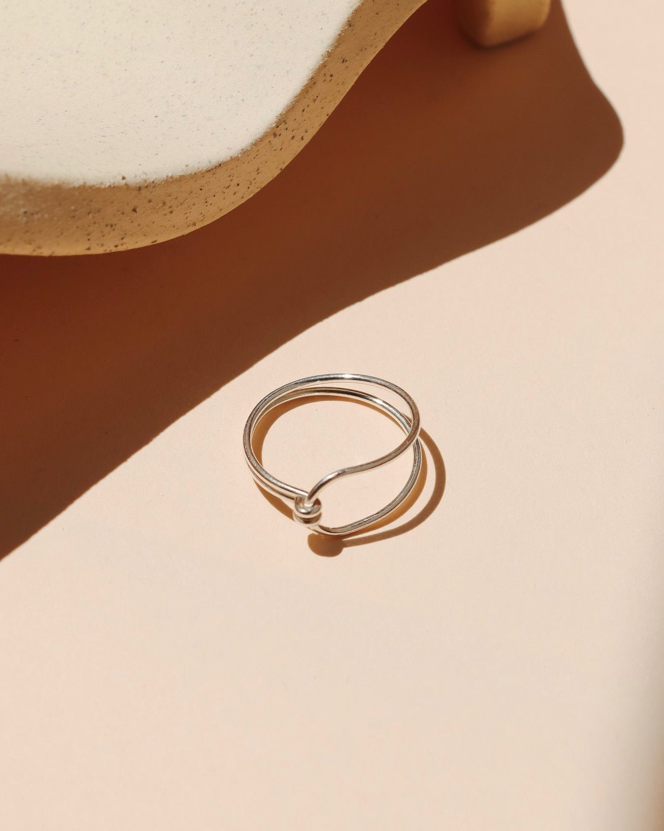 Minimalist fine silver ring with a double-band design joined by a small tied knot detail, set against a warm beige background with soft, architectural shadows and a speckled ceramic prop.