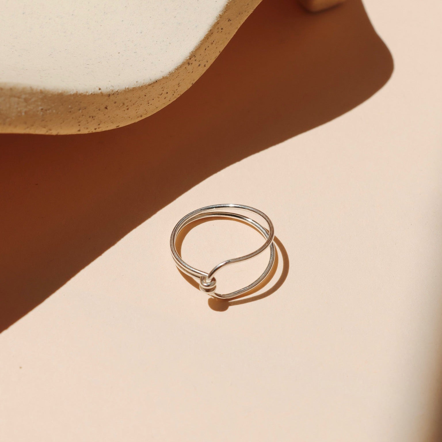 Minimalist fine silver ring with a double-band design joined by a small tied knot detail, set against a warm beige background with soft, architectural shadows and a speckled ceramic prop.