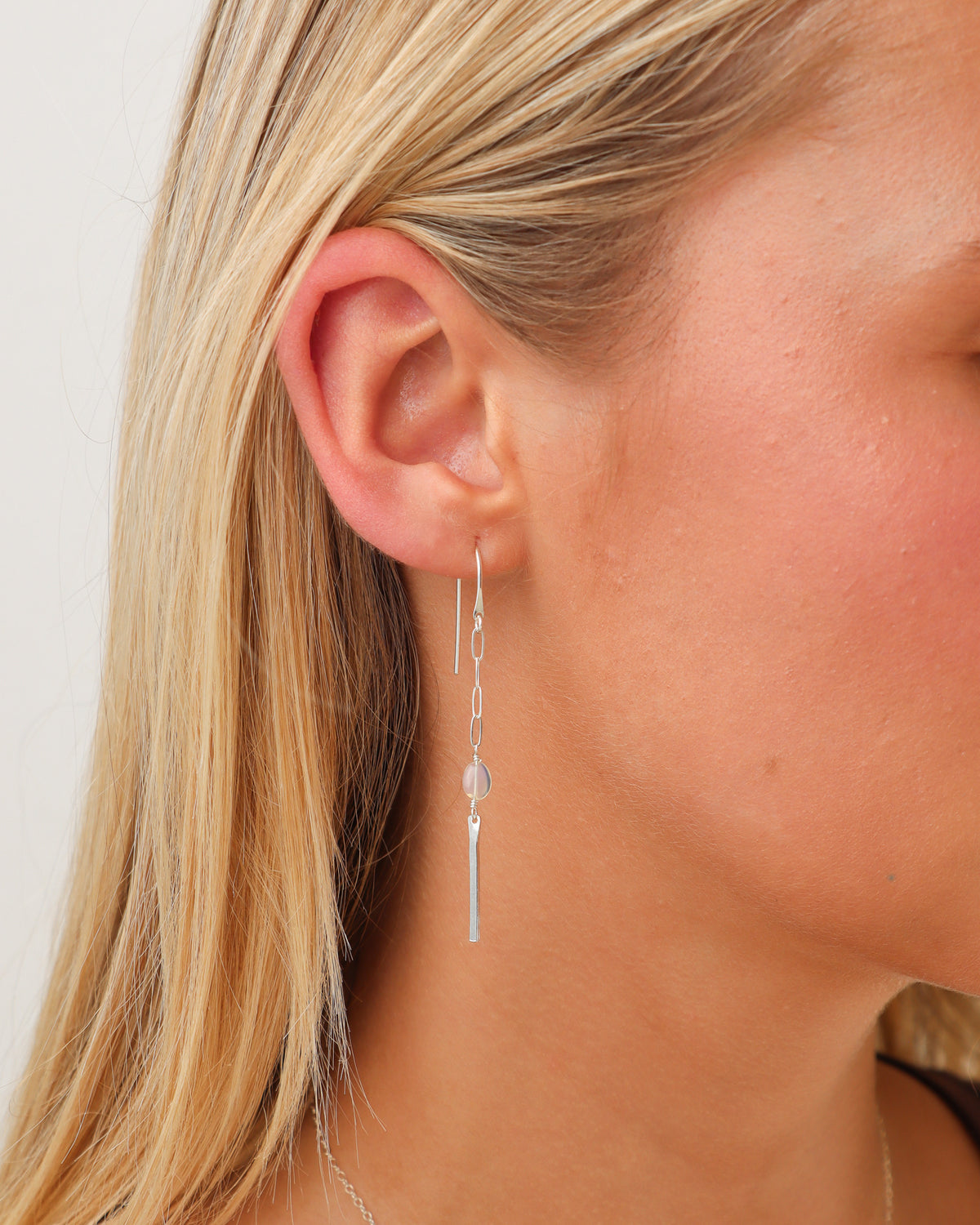 Close-up of a model wearing a silver opal drop earring featuring delicate elongated chain links, an iridescent opal bead, and a slender silver bar pendant against blonde hair.
