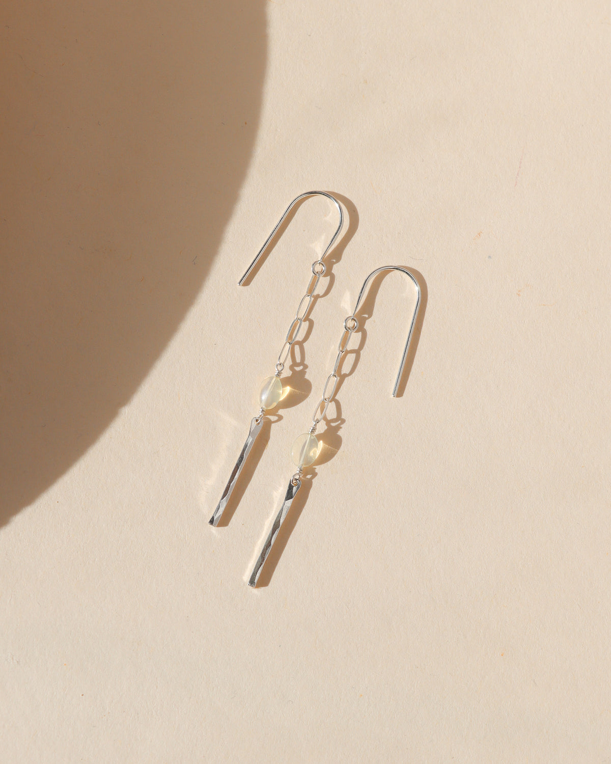 Pair of delicate silver drop earrings featuring elongated chain links, luminous opal beads, and hammered silver bar pendants, arranged on a warm cream background with soft sunlight and shadows.