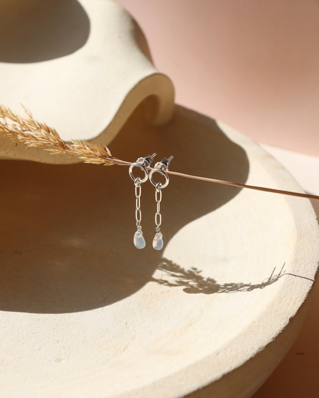 Pair of silver opal drop earrings featuring circular studs, delicate elongated chain links, and iridescent teardrop opal charms, hanging from a dried grass stem above a textured cream ceramic dish in warm sunlight.