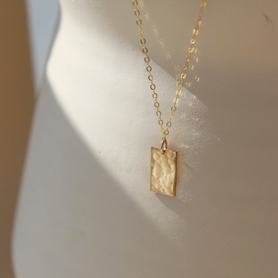 Rectangle 14k gold filled charm suspended from a delicate cable chain with clasp closure. Charm is hammered to a pretty shine and displayed on a white plate. Handmade by Token Jewelry in Eau Claire, WI Box Charm Necklace - Token Jewelry  Edit alt text