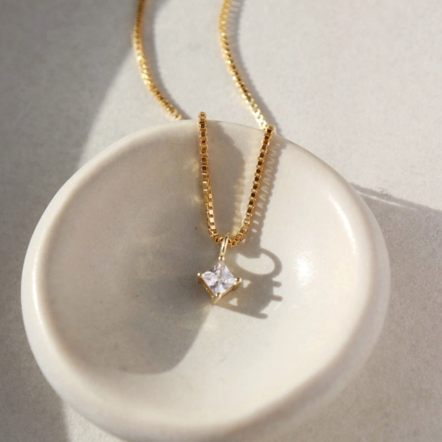 square cut cubic zirconia pendant set in a gold filled bezel and hanging from a delicate box chain sitting on a white jewelry dish.