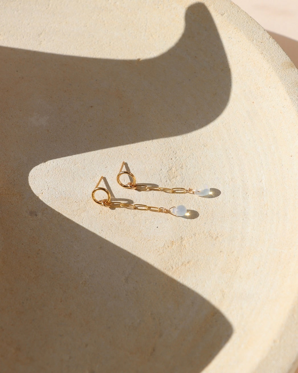 Pair of gold opal drop earrings featuring circular studs, delicate elongated chain links, and iridescent teardrop opal charms, photographed on a textured cream surface in warm sunlight with soft shadows.