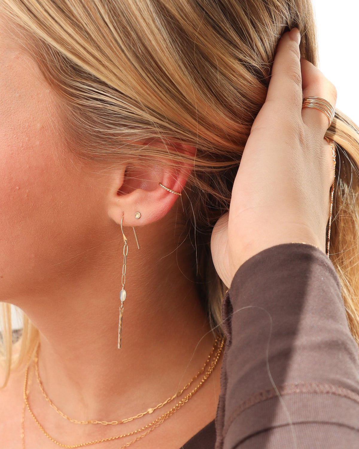 Close-up of a woman wearing a tiny gold stud earring, twisted gold ear cuff, and delicate gold drop earring with a pearl accent, styled with layered gold necklaces and a stacked gold ring.